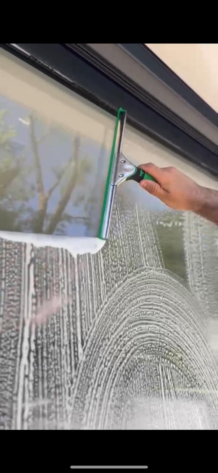 Hand using a squeegee to clean soapy glass window with a reflection of trees.